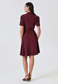 Burgunderfarbenes Hemdkleid mit kurzen Ärmeln, tailliertem Schnitt und ausgestellt Rock. Glattes Material mit einem eleganten Finish und subtiler Satinock.