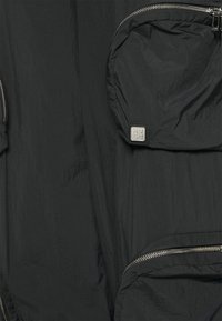 Zwarte jas van een gestructureerde, lichte stof. Voorzien van ritszakken en een klein logo met een glanzende zilveren afwerking.