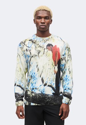 Mann mit kurzen blonden Haaren trägt einen langärmligen Pullover mit dem Aufdruck einer Person in rotem Hemd, die sich vorgebeugt vor einem verschwommenen blau-weißen Hintergrund befindet.