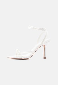 Λευκό σανδάλι με ψηλό τακούνι, λεπτά λουράκια στον αστράγαλο και τα δάχτυλα, διακοσμημένα με μικρές πέρλες, και κούμπωμα με πόρπη.