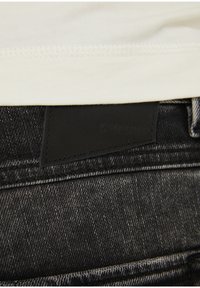 Jeans negros con un acabado texturizado, que cuentan con una etiqueta de cuero negro liso cosida en la cinturilla que muestra el nombre de la marca.