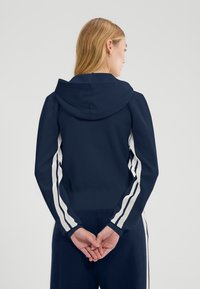 Felpa con cappuccio blu navy con dettagli a strisce bianche lungo le maniche, trama a maglia e polsini e orlo a coste. Vista posteriore che mostra le caratteristiche del design.