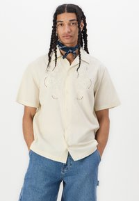 Camisa beige de manga corta con bordado floral, cierre de botones y un corte relajado, combinada con shorts de mezclilla azules y un pañuelo azul.