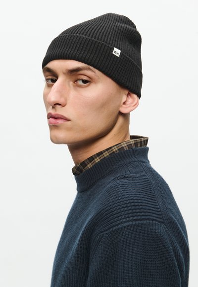 Crna rebrasta kapa s malim bijelim logotipom, nosi je osoba u tamnoplavom teksturiranom puloveru s kragnom na karirane košulje ispod.