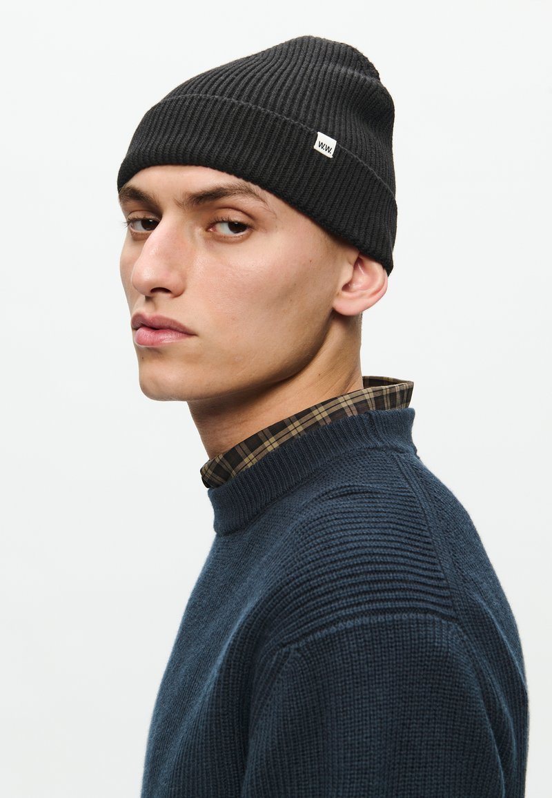Black ribbed beanie with a small white logo, worn by a person in a dark blue textured sweater with a plaid shirt collar underneath.
