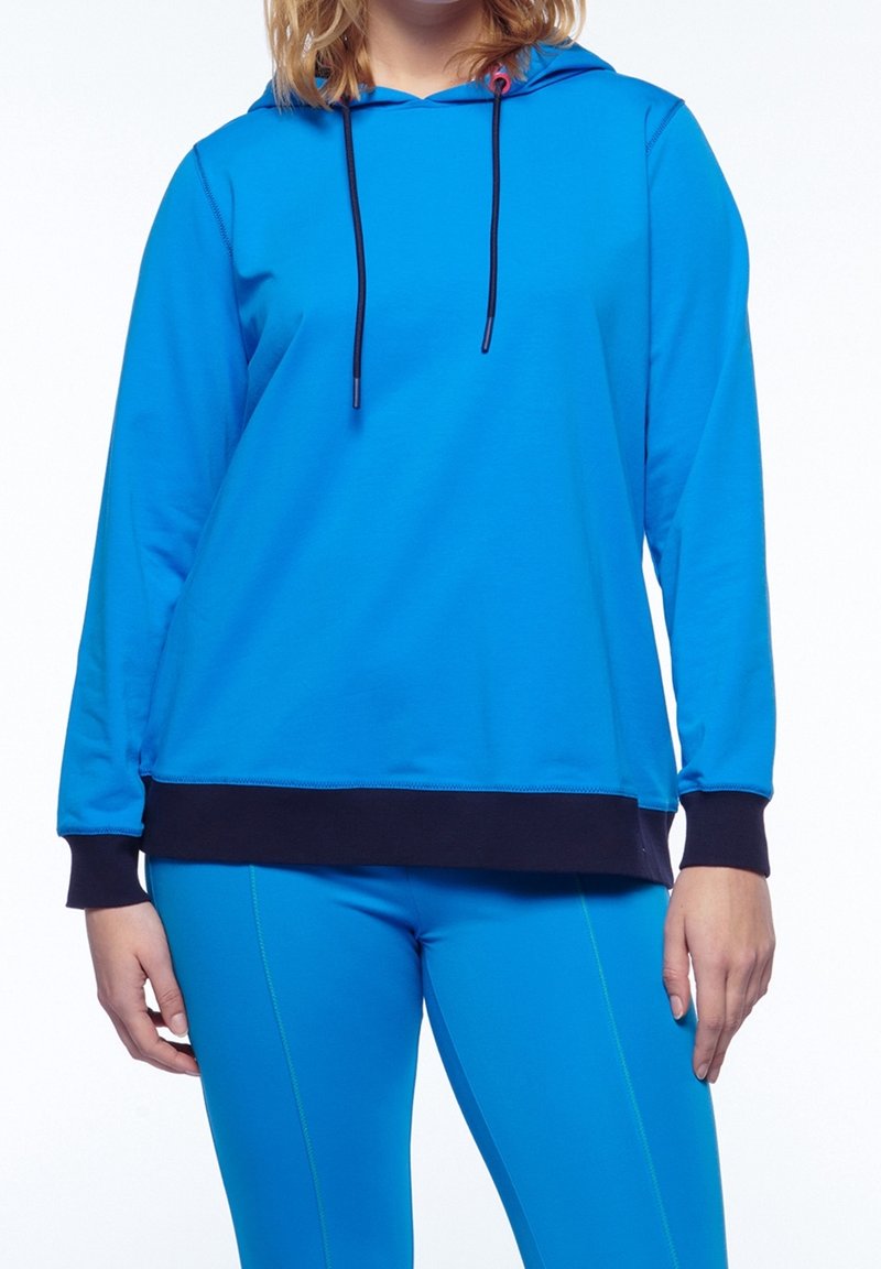 Femme portant un sweat à capuche bleu vif avec des poignets noirs et un pantalon assorti bleu ajusté, debout sur un fond blanc.