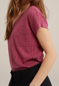 Pletený top v magentové barvě s výstřihem do V, krátkými rukávy a otevřeným vzorem stehu. Kombinovaný s černými kalhotami s vysokým pasem.