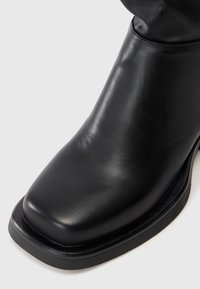 Bota de tornozelo em couro preto com bico quadrado e salto baixo num fundo branco.