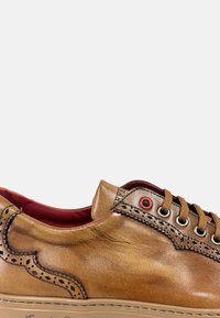 Zapatilla de cuero marrón con intrincados bordados perforados, forro rojo y ojales metálicos. Punta redondeada y detalle de suela en contraste.