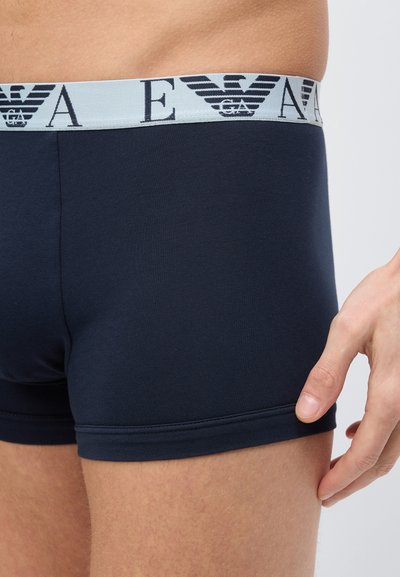 Κοντινό πλάνο ενός ατόμου που φοράει σκούρο μπλε ανδρικά εσώρουχα boxer με γκρι ζώνη μέσης που φέρει μαύρο λογότυπο και γράμματα, με το χέρι να ακουμπάει κοντά στο μηρό.