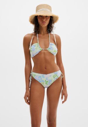 Kobieta w pastelowym kwiatowym bikini z wiązaniami po bokach oraz szerokim kapeluszu słomkowym z paskami "MALINA", stojąca na białym tle.