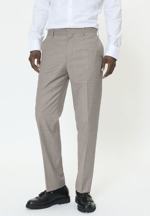 Mand iført lysebrune, skræddersyede bukser, hvid skjorte og sorte loafers med kvaster, stående mod en ensartet hvid baggrund.