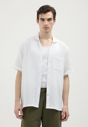 Giovane uomo che indossa una camicia bianca a maniche corte con bottoni sopra una canottiera bianca, abbinata a pantaloni con coulisse verde oliva, in posizione neutra.