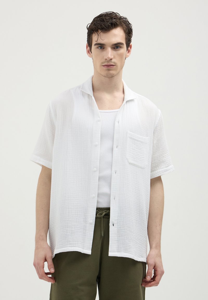Giovane uomo che indossa una camicia bianca a maniche corte con bottoni sopra una canottiera bianca, abbinata a pantaloni con coulisse verde oliva, in posizione neutra.