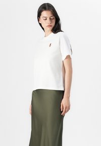 Modell med långt mörkt hår bär en vit t-shirt med en liten logotyp och en grön satin kjol, stående mot en enkel vit bakgrund.