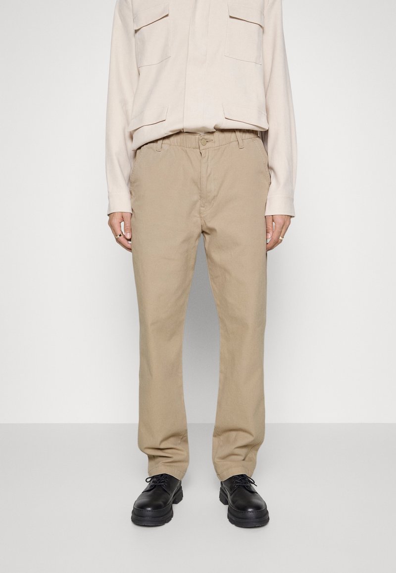 Personne portant un pantalon beige droit, une chemise beige clair à manches longues et des chaussures noires à lacets, debout devant un fond uni.