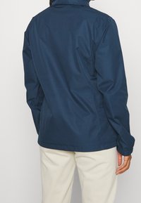 Personne portant une veste bleu marine avec des poignets élastiques et un pantalon beige, vue de dos sur un fond uni.