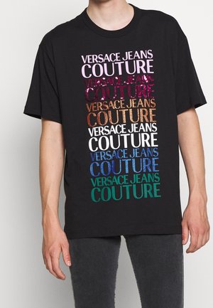 Czarny T-shirt z kolorowym, błyszczącym napisem "VERSACE JEANS COUTURE" w różnych rozmiarach i czcionkach. Krótkie rękawy, okrągły dekolt.