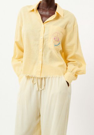 Henkilö, jolla on päällään löysä keltainen nappipaita, jossa on abstrakti kasvojen kirjailu taskussa, ja vaalean kermaan vivahtavat nauhalliset housut, kädet taskuissa.