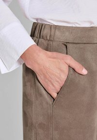 Bruine suède broek met een losse pasvorm, voorzien van zijzakken en een elastische tailleband. Een witte blouse met lange mouwen is gedeeltelijk zichtbaar.
