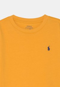 Gelbes Baumwoll-T-Shirt mit Rundhalsausschnitt, kurzen Ärmeln und einem kleinen blauen gestickten Polo-Logo auf der linken Brust. Glatte Textur.