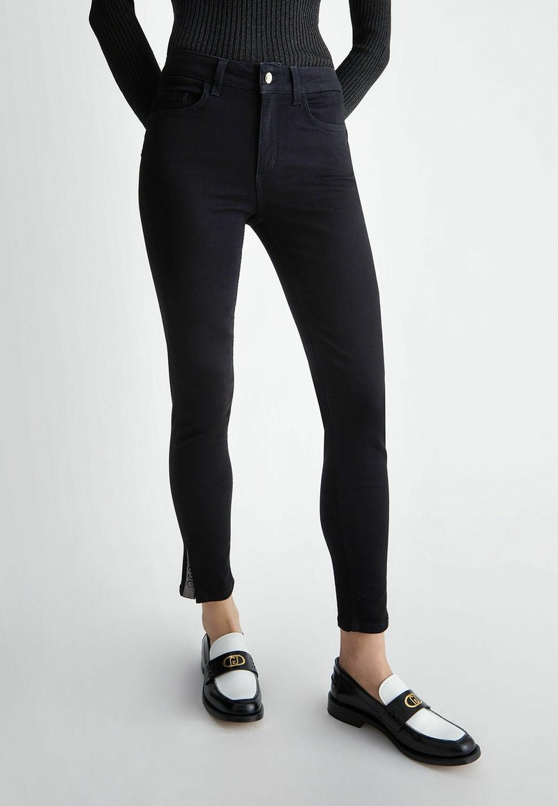 Jean skinny noir taille haute en tissu texturé avec un subtil détail en forme de découpe au niveau de la cheville, associés à des mocassins noirs et blancs.