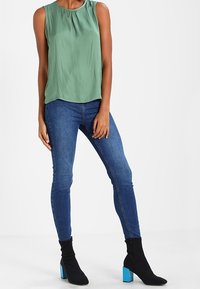 Zelený top bez rukávů s kulatým výstřihem, spárovaný s modrými skinny džíny a černými kotníkovými botami s modrými blokovými podpatky.