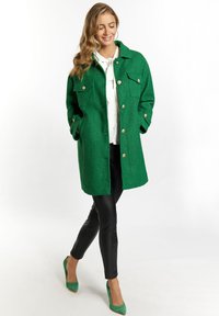 Cappotto verde con texture, dotato di grandi tasche e bottoni dorati, indossato sopra una blusa bianca, abbinato a leggings neri e tacchi verdi.