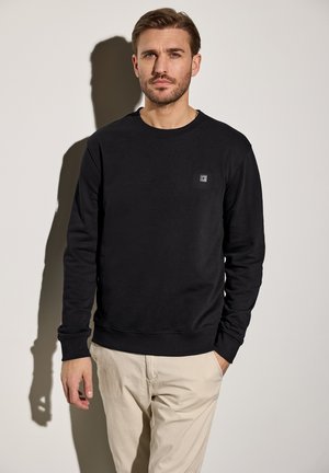 Zwarte crewneck sweatshirt gemaakt van zacht materiaal met ribbelmanchetten en -zoom. Bevat een klein logopatch op de borst. Lichte achtergrond.