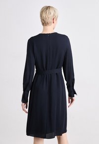 Jūras zila apģērbs ar garām piedurknēm, jostas saiti un aizmugurējo rāvējslēdzēju. Gluda auduma izstrādājums ar ceļa garuma apakšmalu un smalku šķēlumu detaļu pie piedurknēm.