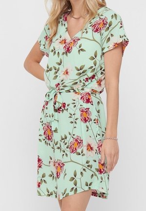 Mujer con un vestido cruzado de manga corta color verde claro con un patrón floral en rojo, rosa y amarillo, atado a la cintura con un cinturón a juego.