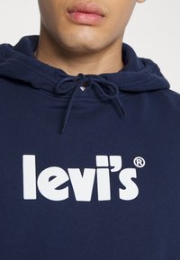 Námořnicky modrá mikina s kapucí a stahovací šňůrkou, na přední straně s výrazným bílým logem "levi's". Měkký bavlněný materiál, ležérní design.