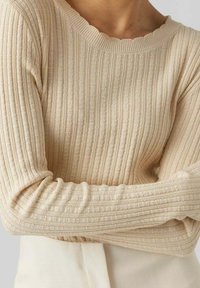 Pull en tricot beige côtelé avec des motifs en torsades, porté par une personne avec les bras croisés, associé à un pantalon crème taille haute.