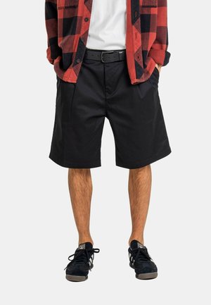 Hombre con pantalones cortos negros hasta la rodilla, cinturón negro, zapatillas negras, camiseta blanca y camisa de cuadros roja y negra con las manos en los bolsillos.