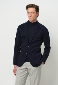 Blazer navy realizzato in tessuto testurizzato, con rever a punta e due tasche frontali, abbinato a un dolcevita scuro e pantaloni grigi chiari.