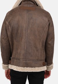 Hnědá bunda s vlněným podšívkou a texturovaným koženým povrchem, se vlněným límcem, bočními přezkami a hladkým látkovým znakem na zadní straně.