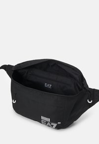 Borsa a spalla in tessuto nero con logo di marca. L'interno presenta una tasca con zip, una tracolla regolabile e una finitura liscia. Si apre dall'alto.