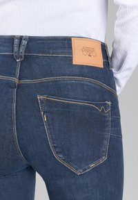 Jeans de mezclilla de un azul oscuro con un diseño de tiro medio, costuras naranjas, un parche de cuero de la marca en la parte posterior y dos bolsillos traseros.