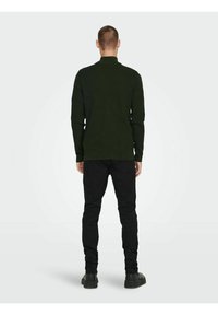 Dark green long-sleeve top with a high collar, paired with black slim-fit pants and black footwear, viewed from the back.