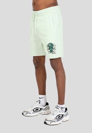 Maschio che indossa pantaloncini verde chiaro con coulisse con una grafica di bambola voodoo in stile cartone animato sulla parte anteriore della gamba sinistra e scarpe da ginnastica Nike bianche.