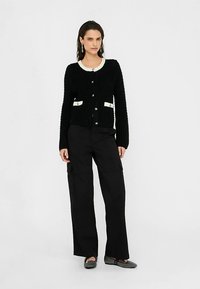 Schwarzer, strukturierter Cardigan mit cremefarbenen Akzenten, vorderen Knöpfen und Taschen. Modell trägt schwarze, weit geschnittene Hosen und flache Schuhe.