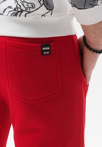 Røde sweatpants har en baglomme med et sort label, der står "STAY REAL, SINCE 1995." Stoffet ser blødt og tekstureret ud.