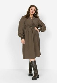 Vestido a rayas marrón y beige con un escote abotonado, cintura fruncida y mangas largas abullonadas. Combinado con botas marrones hasta la rodilla.