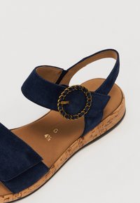 Granatowy zamszowy sandał z korkową podeszwą, wyposażony w szeroki pasek i okrągłą klamrę z łańcuchem w złotym odcieniu do zapięcia.