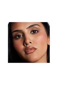 Porträtt av en person med medium hudton, mörkt hår, fylliga läppar med glansig nude läppfärg, väl definierade ögonbryn och subtil ögonmakeup.