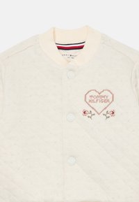 Gepolsterte cremefarbene Bomberjacke mit einem gerippten Kragen, ausgestattet mit gesticktem "Tommy Hilfiger" Herzdesign und floralen Akzenten, gesichert durch Druckknöpfe.