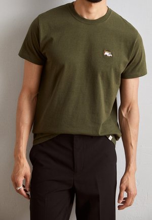 Mężczyzna ubrany w oliwkowozieloną koszulkę z okrągłym dekoltem z małym, haftowanym białym vanem na piersi oraz czarne spodnie, stojący na tle szarej ściany.