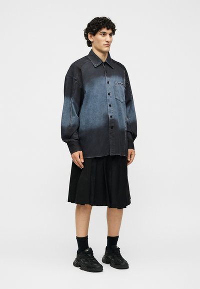 Νέος άνδρας που φορά χαλαρό ξεθωριασμένο μαύρο πουκάμισο με κουμπιά, μαύρη πλισέ φούστα, μαύρες κάλτσες και μαύρα αθλητικά παπούτσια, στέκεται μπροστά από ένα απλό φόντο.