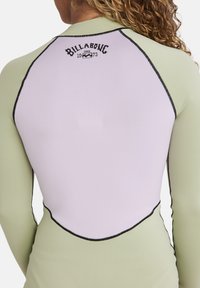Maglia a maniche lunghe in verde chiaro e bianco. Presenta cuciture nere e un logo in stile vintage sul retro. Tessuto elastico e liscio.