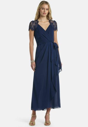 Kobieta ubrana w granatową sukienkę typu wrap z koronkowymi, krótkimi rękawami, ozdobioną boczną kokardą, w złotych sandałach na wysokim obcasie, stojąca na białym tle.